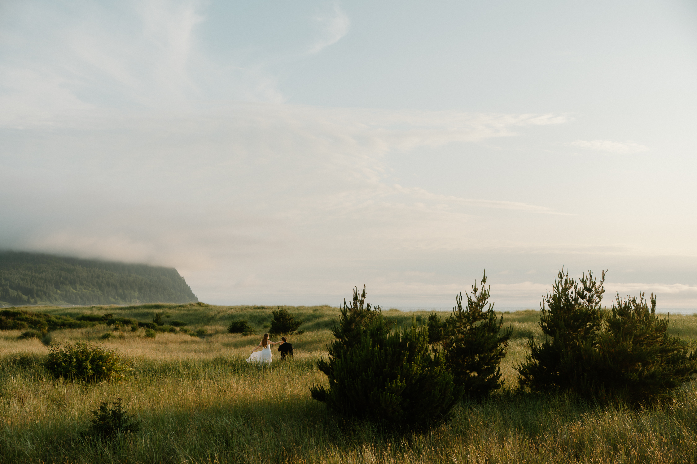 A couple plays in tall green grass with a mountain backdrop during their Oregon Coast elopement. 