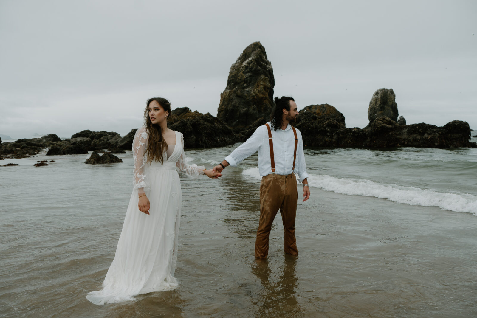 A couple stands in the ocean in front of black rocks at Cannon Beach during their elopement. 