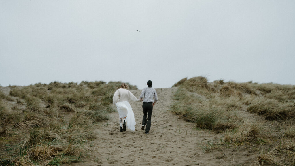 A couple runs towards the beach between grassy dunes as the wind catches their clothes and hair during their Cannon Beach engagement photo session. 