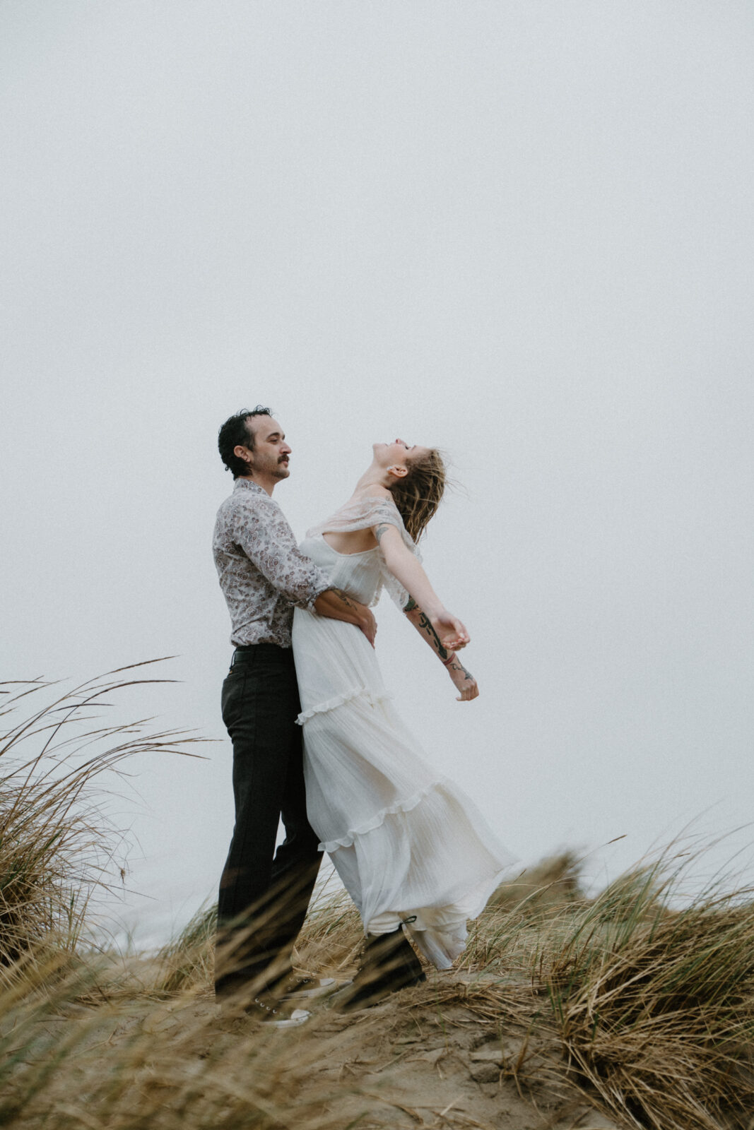 A couple stands high on a grassy dune and embraces as the wind whips at them during their Cannon Beach engagement photo session