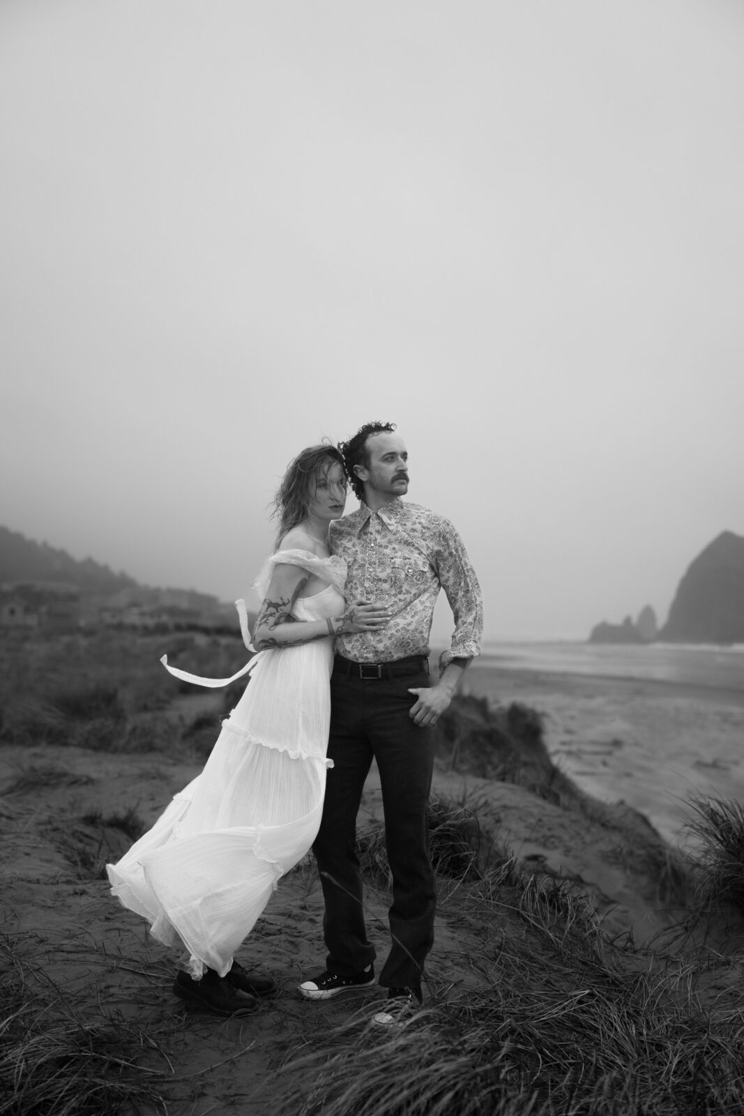 A black and white photo of a couple standing on a grassy dune, with Haystack rock in the background, during their Cannon Beach engagement photo session. 