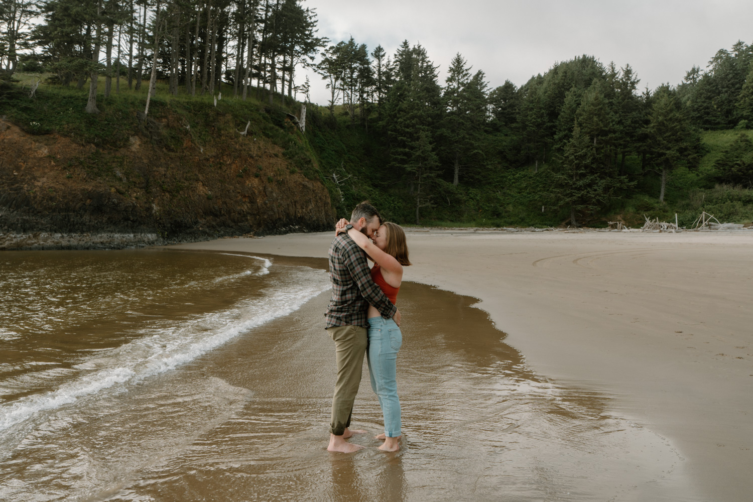 A couple stands in the ocean in front of a forested cliff for their Cannon Beach engagement photos. 