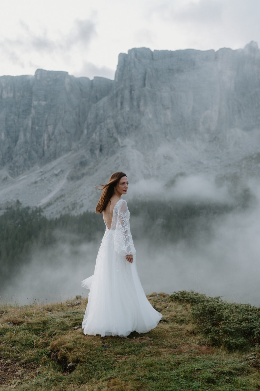 A bride stands in a grass field with a moody, cloudy, mountain scene behind her during her Lago di Braies elopement. 
