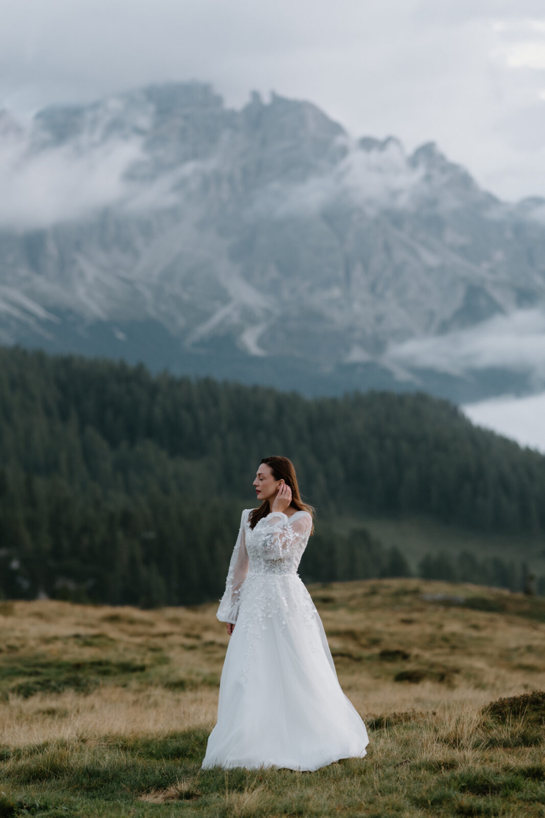 A bride stands in a grass field with a moody, cloudy, mountain scene behind her during her Lago di Braies elopement. 