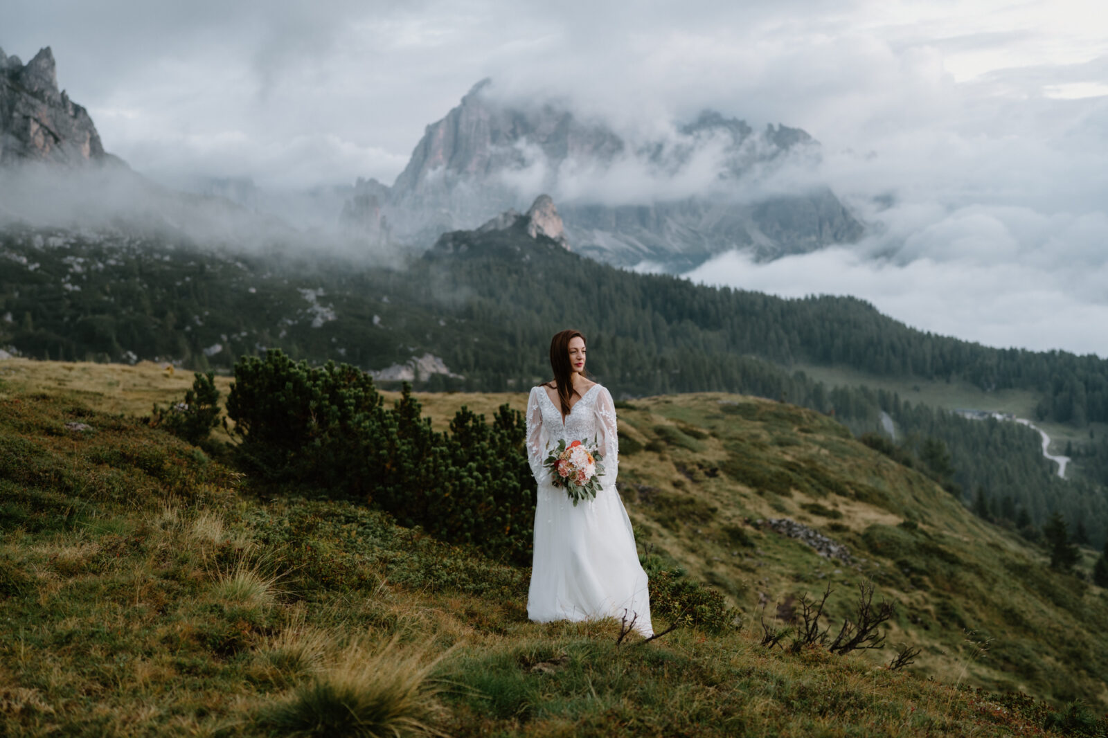 A bride stands in a grass field with a moody, cloudy, mountain scene behind her during her Lago di Braies elopement. 