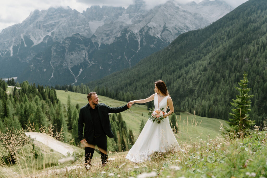 A couple stands in an alpine meadow with flowers and tall, cloudy mountains in the background during their Lago di Braies elopement. 