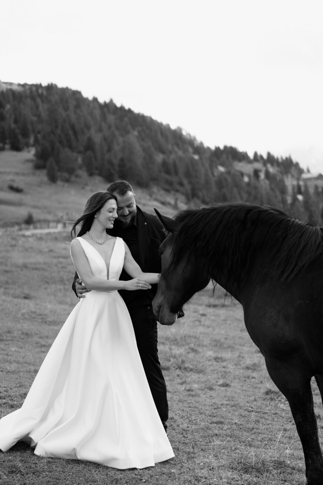 A couple in wedding attire snuggle together and reach for a black horse during their Lago di Braies elopement. 