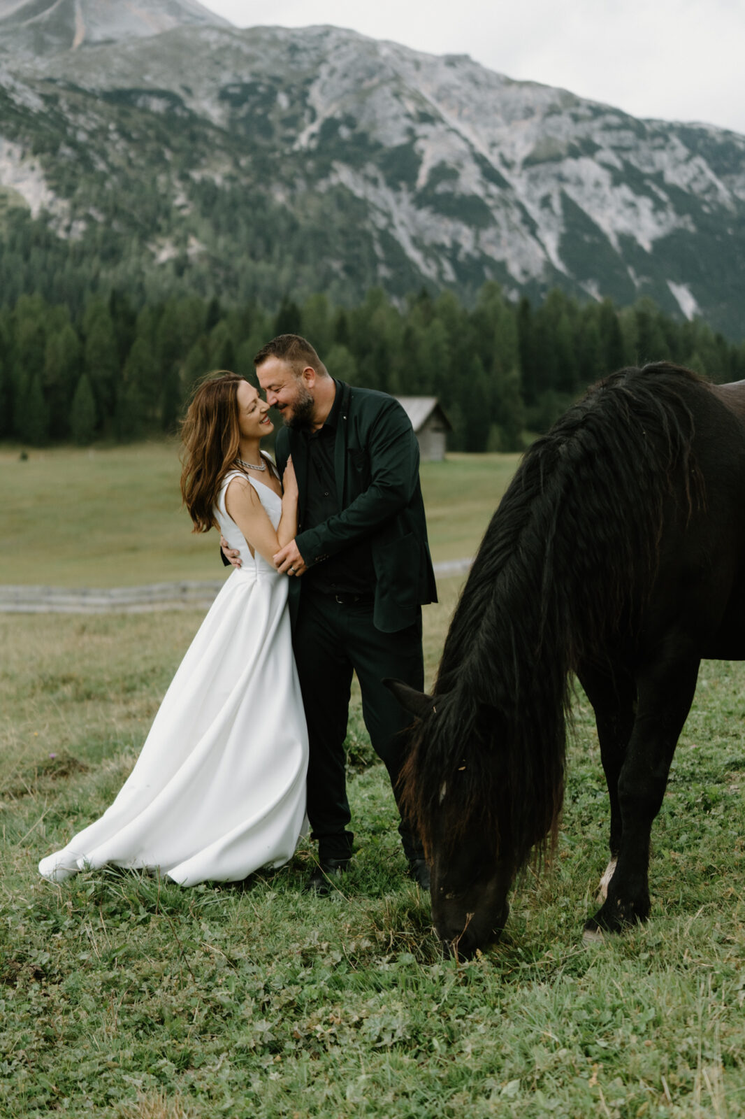 A couple in wedding attire snuggle together and reach for a black horse during their Lago di Braies elopement. 