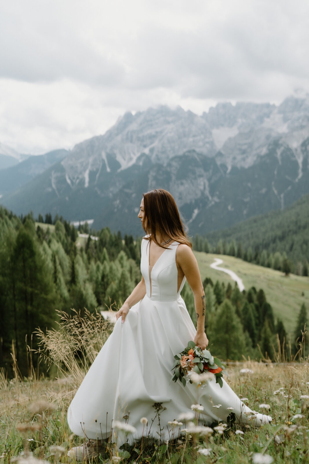 A bride with a bouquet walks throw a flowery alpine meadow in front of cloudy mountains during her Lago di Braies elopement. 