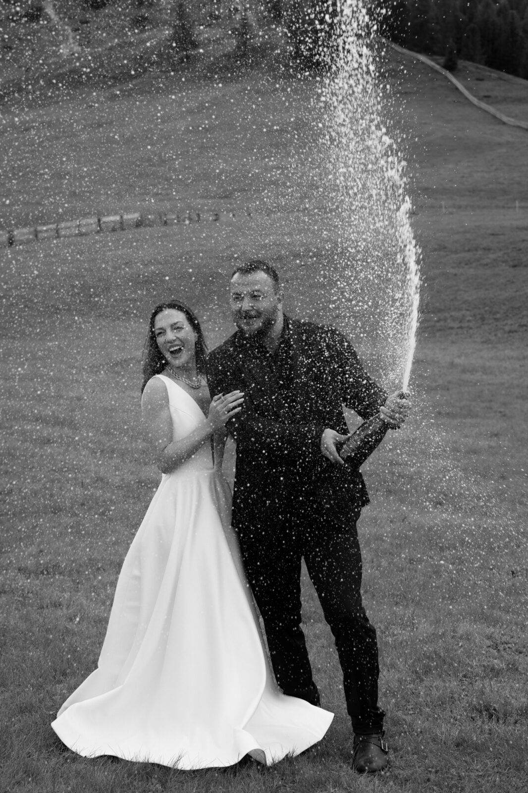 A couple in wedding attire stands in an alpine field and sprays champagne to celebrate their Lago di Braies elopement. 