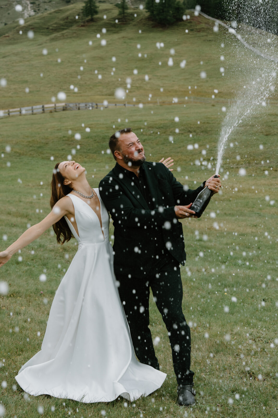 A couple in wedding attire stands in an alpine field and sprays champagne to celebrate their Lago di Braies elopement. 