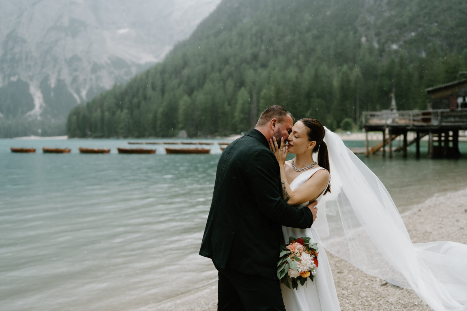A Lago di Braies elopement photo, with a groom and bride kissing in front of the lake while the rain comes down around them. 