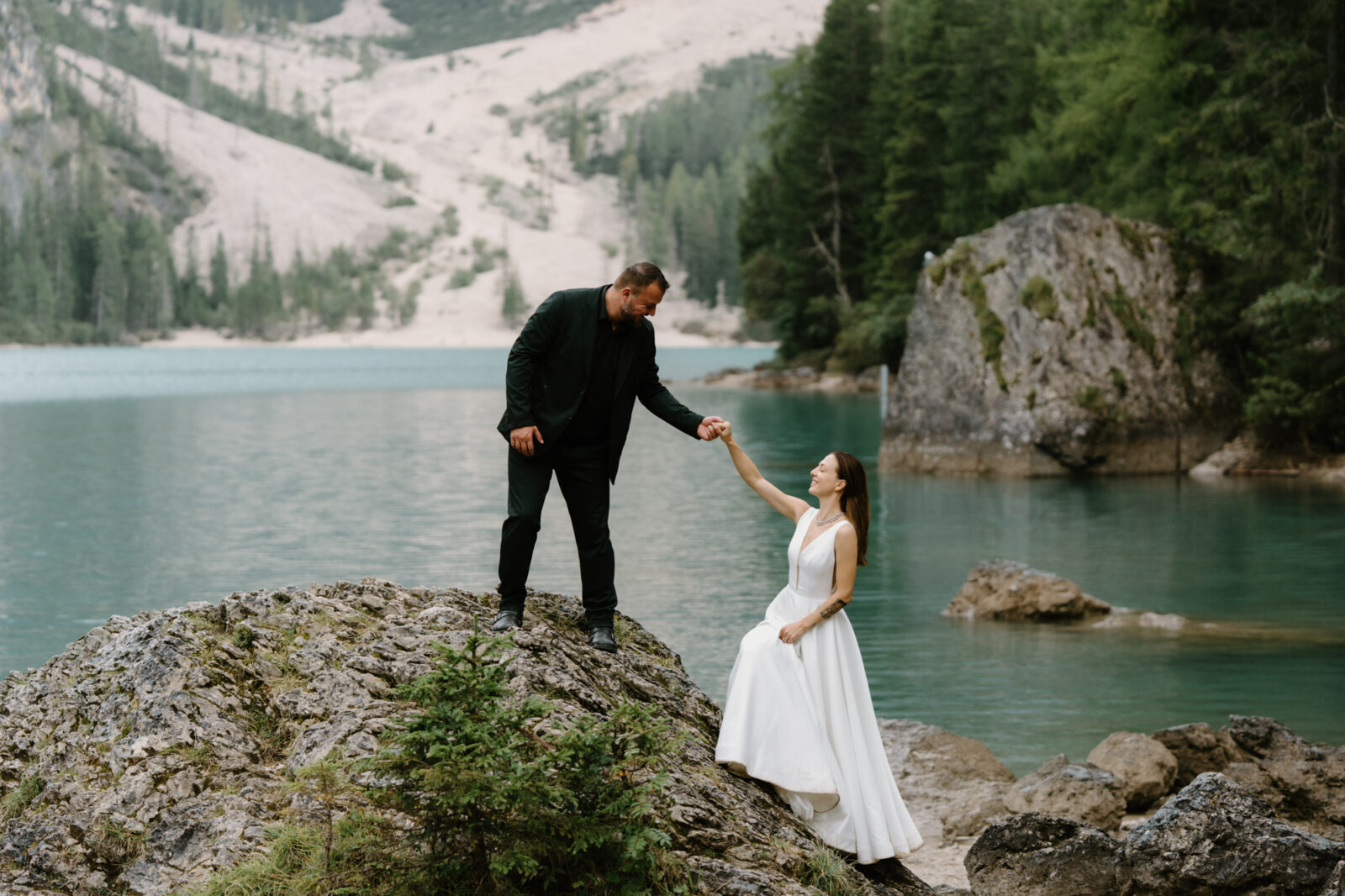 A groom helps a bride up onto a tall rock in front of an alpine lake during their Lago di Braies elopement. 