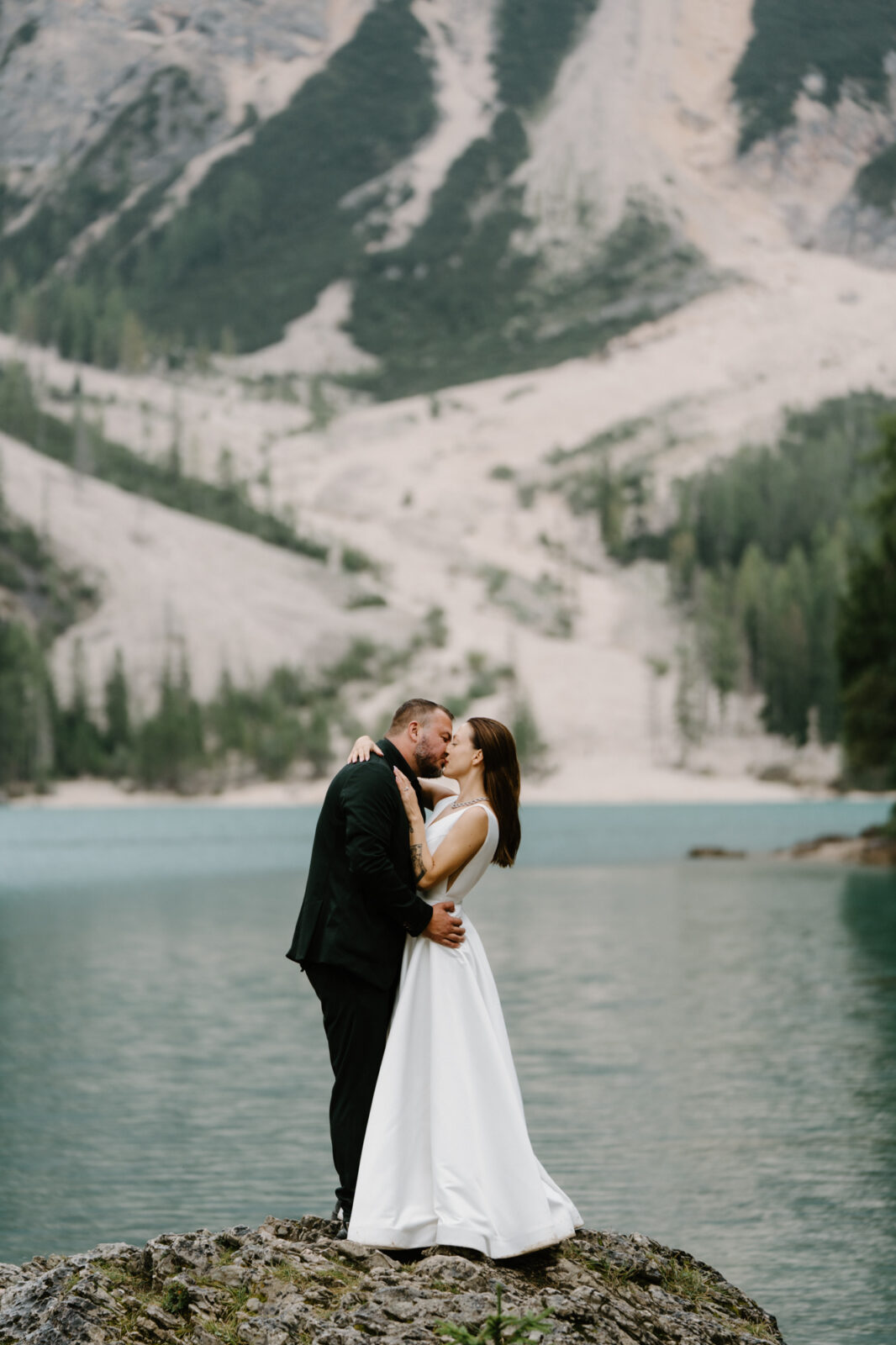 A couple in wedding attire standing on a rock in front of a turquoise alpine lake during their Lago di Braies elopement.  