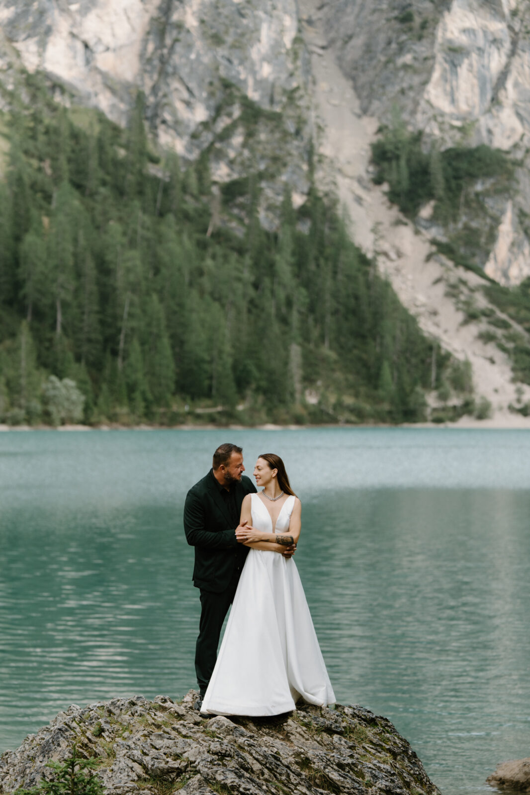 A couple in wedding attire standing on a rock in front of a turquoise alpine lake during their Lago di Braies elopement.  