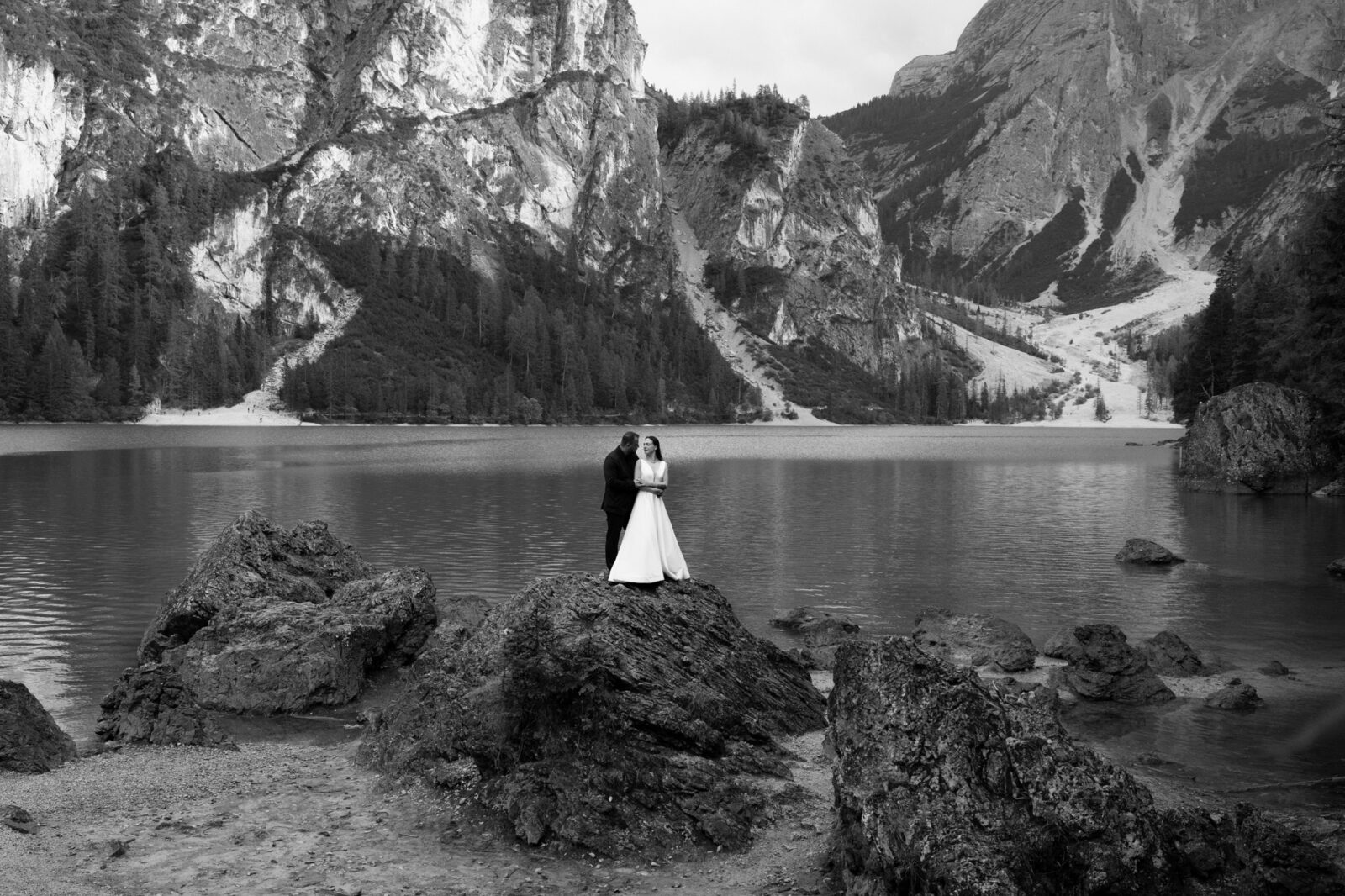 A black and white photo of a couple stand on a rock, far in the distance, with an alpine lake behind them for their A couple in wedding attire standing on a rock in front of a turquoise alpine lake during their Lago di Braies elopement.  