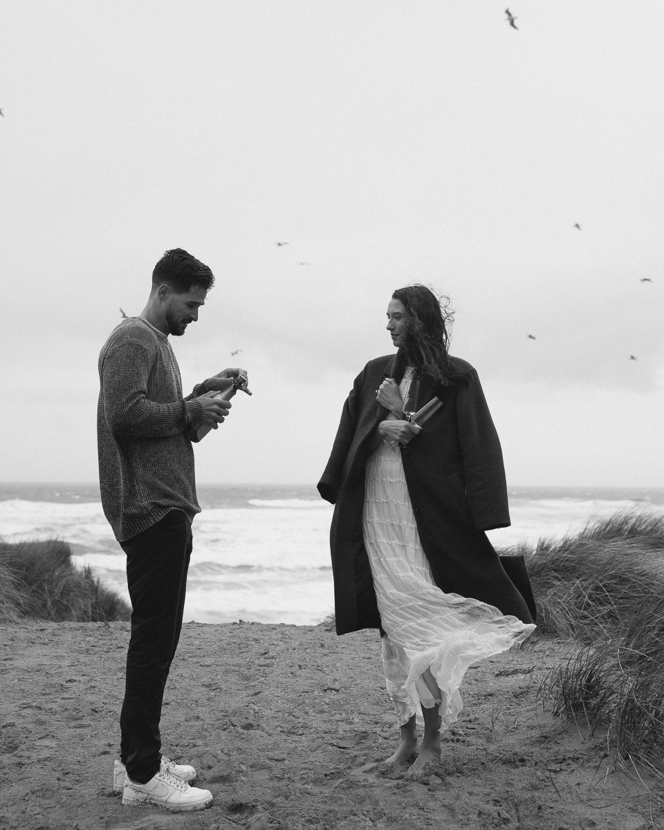 A couple pops champagne at their Oregon coast engagement photo shoot.