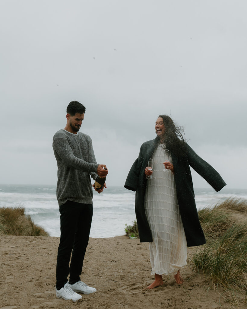 A couple pops champagne at their Oregon coast engagement photo shoot.