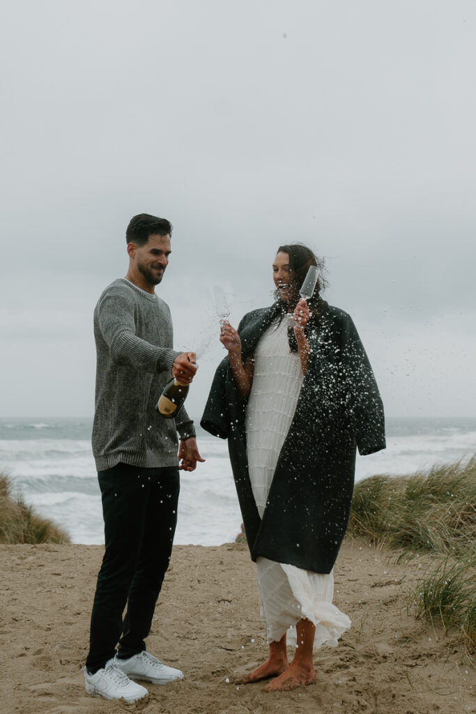 A couple pops champagne at their Oregon coast engagement photo shoot.