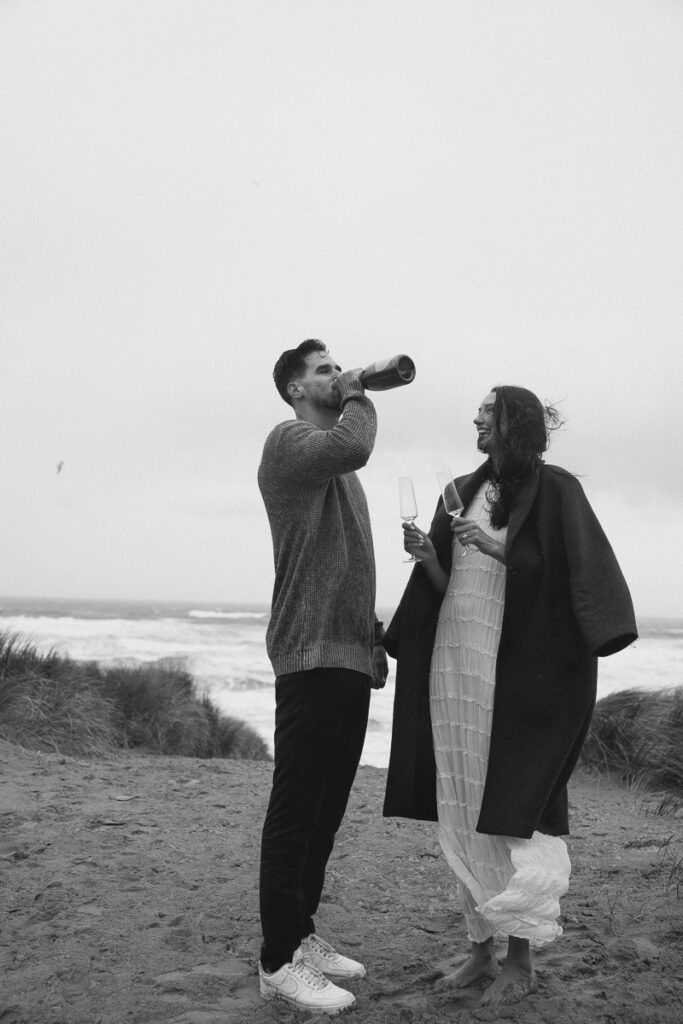 A couple pops champagne at their Oregon coast engagement photo shoot.
