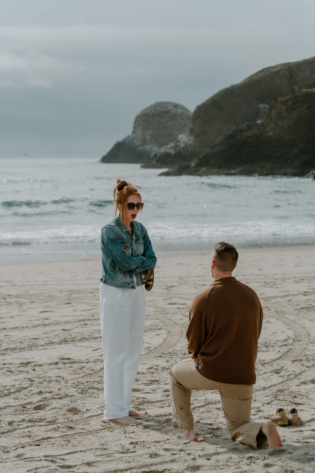 A woman stands with a surprised look on her face while her boyfriend is down on one knee for an Oregon coast proposal.