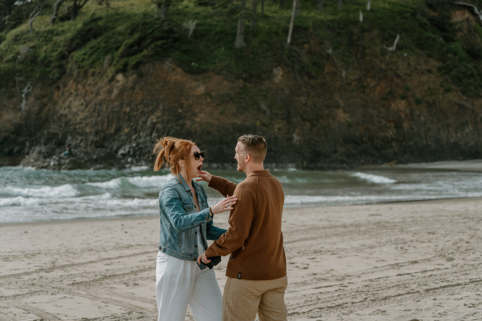 A woman stands with a surprised look on her face while her boyfriend is down on one knee for an Oregon coast proposal.