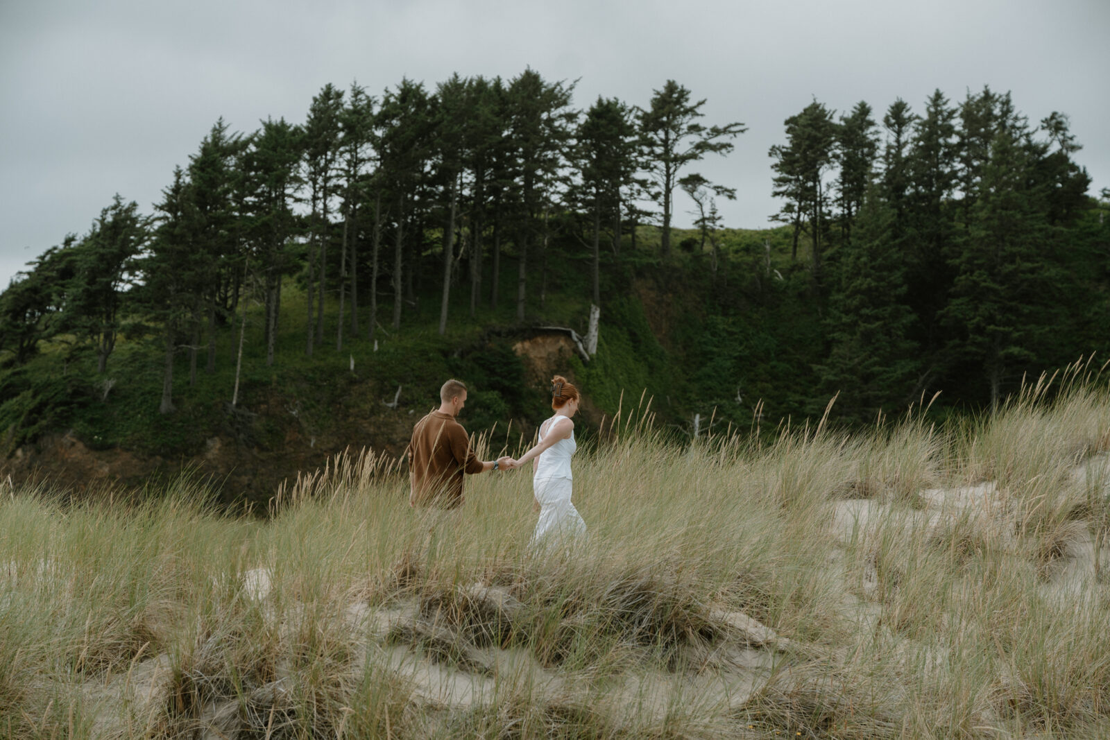 A couple walks through tall seagrass in front of a rock with dark green trees after their Oregon coast proposal session.