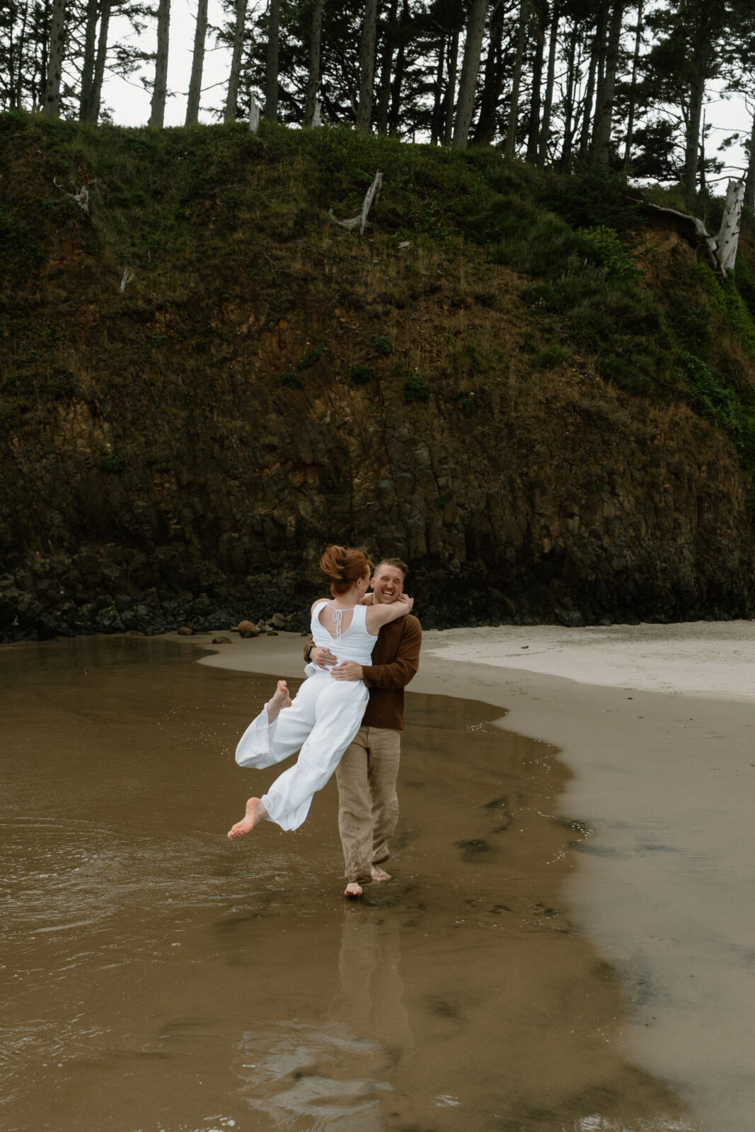 A man and woman walk together and laughs as they splash in the ocean after their Oregon Coast proposal.