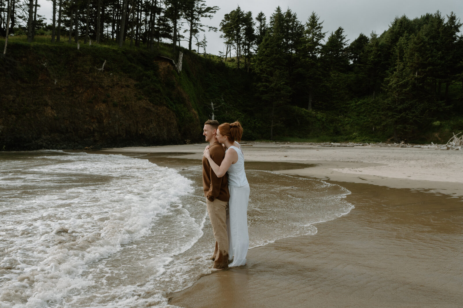 A couple stands in the water and looks out at the ocean after their Oregon Coast proposal.