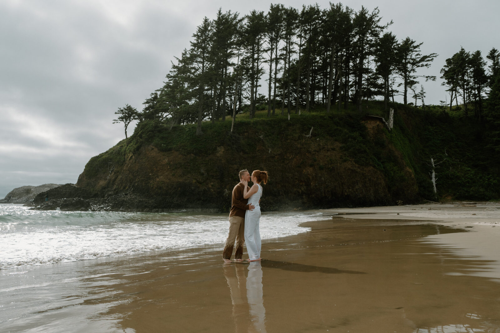 A couple stands on the beach in front of a rock and trees and kisses at sunset.