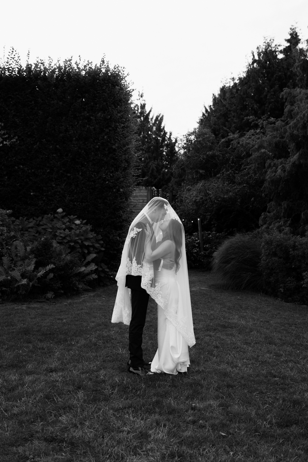 A couple in wedding attire poses under a veil in black and white at their Briarwood Estate wedding.