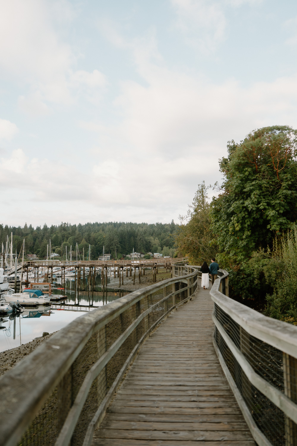 A couple walks along a harbor with boats on Bainbridge island while they have their seattle ferry engagement photos taken.