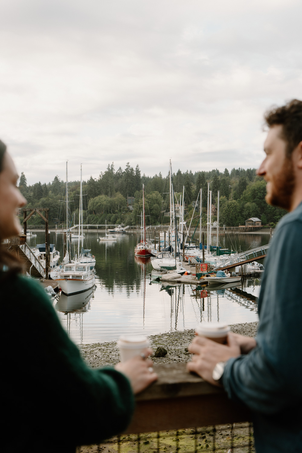 A couple walks along a harbor with boats on Bainbridge island while they have their seattle ferry engagement photos taken.