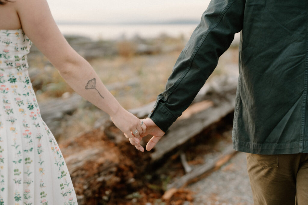 A couple holds hands on a rocky beach on bainbridge island during their seattle ferry engagement photos session.
