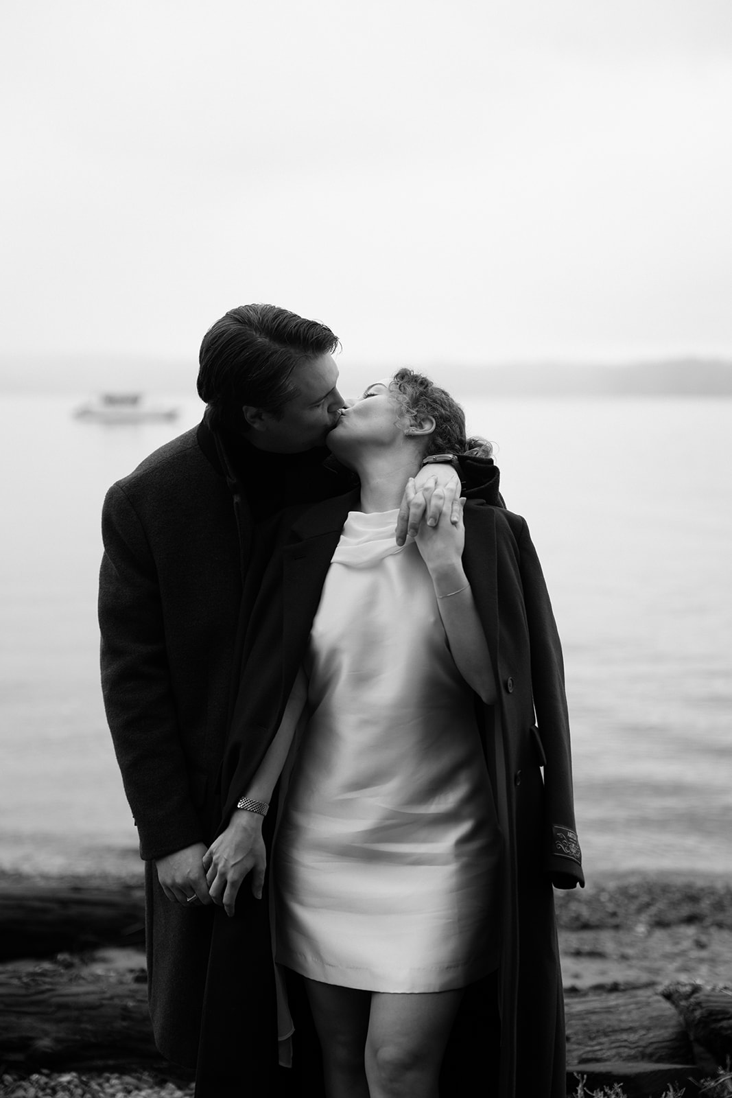 A black and white photo of a couple snuggling together on a shoreline in front of a ferry for their Seattle engagement photos