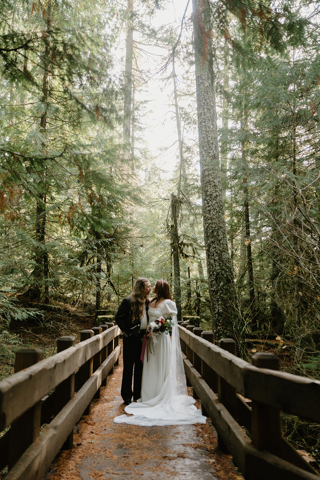 A couple in wedding attire stands in the middle of a tall forest during their Sahalie Falls elopement. 