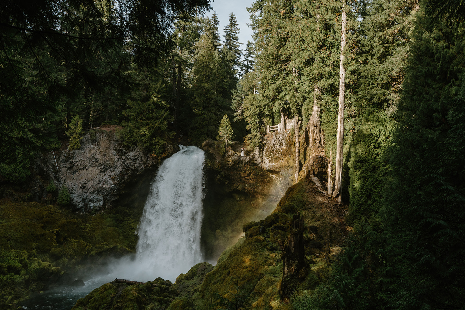 A couple in wedding attire stands small in front of a giant waterfall during their Sahalie Falls elopement. 