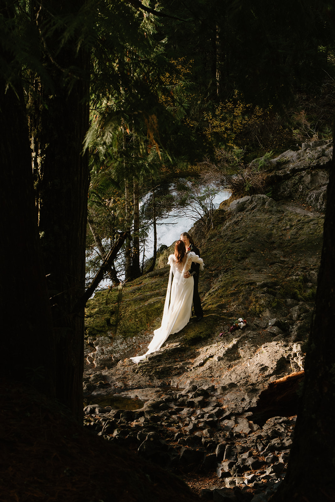 A couple in wedding attire stands, facing the camera, at the top of a waterfall with a misty forest behind them during their Sahalie Falls elopement. 