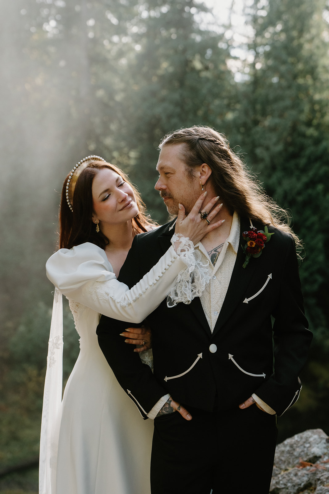 A couple in wedding attire stands, facing the camera, at the top of a waterfall with a misty forest behind them during their Sahalie Falls elopement. 