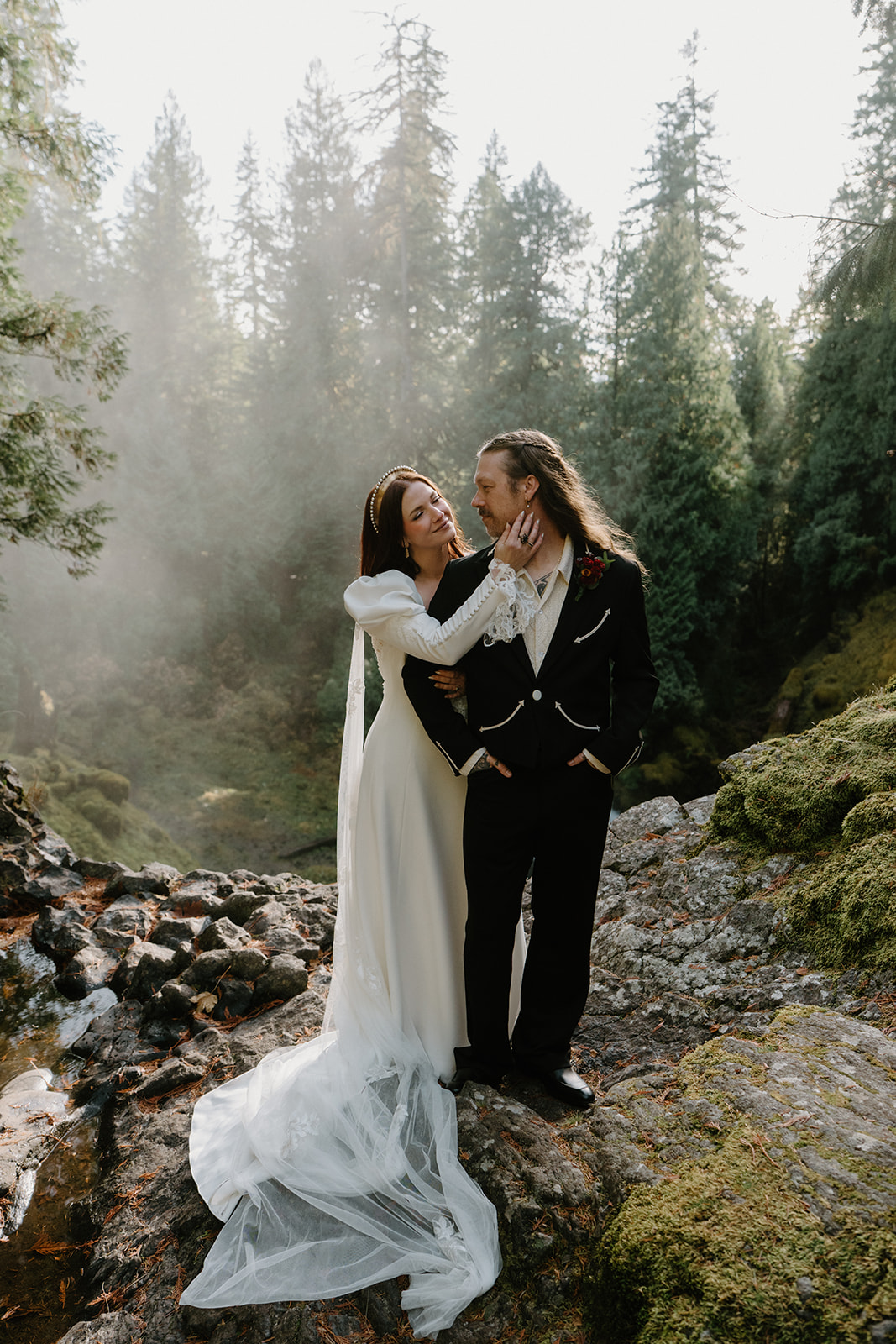 A couple in wedding attire stands, facing the camera, at the top of a waterfall with a misty forest behind them during their Sahalie Falls elopement. 