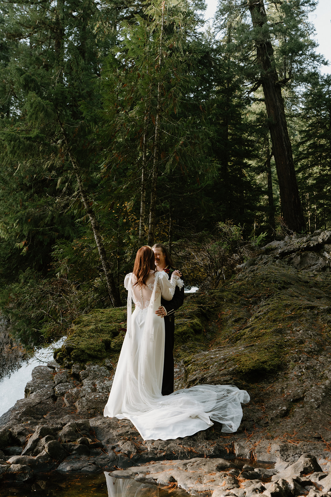 A couple in wedding attire stands, facing the camera, at the top of a waterfall with a misty forest behind them during their Sahalie Falls elopement. 
