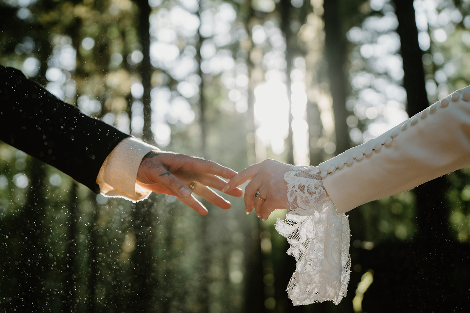 A couple in wedding attire reach for each others hands as mist sparkles around them in the forest during their Sahalie Falls elopement. 