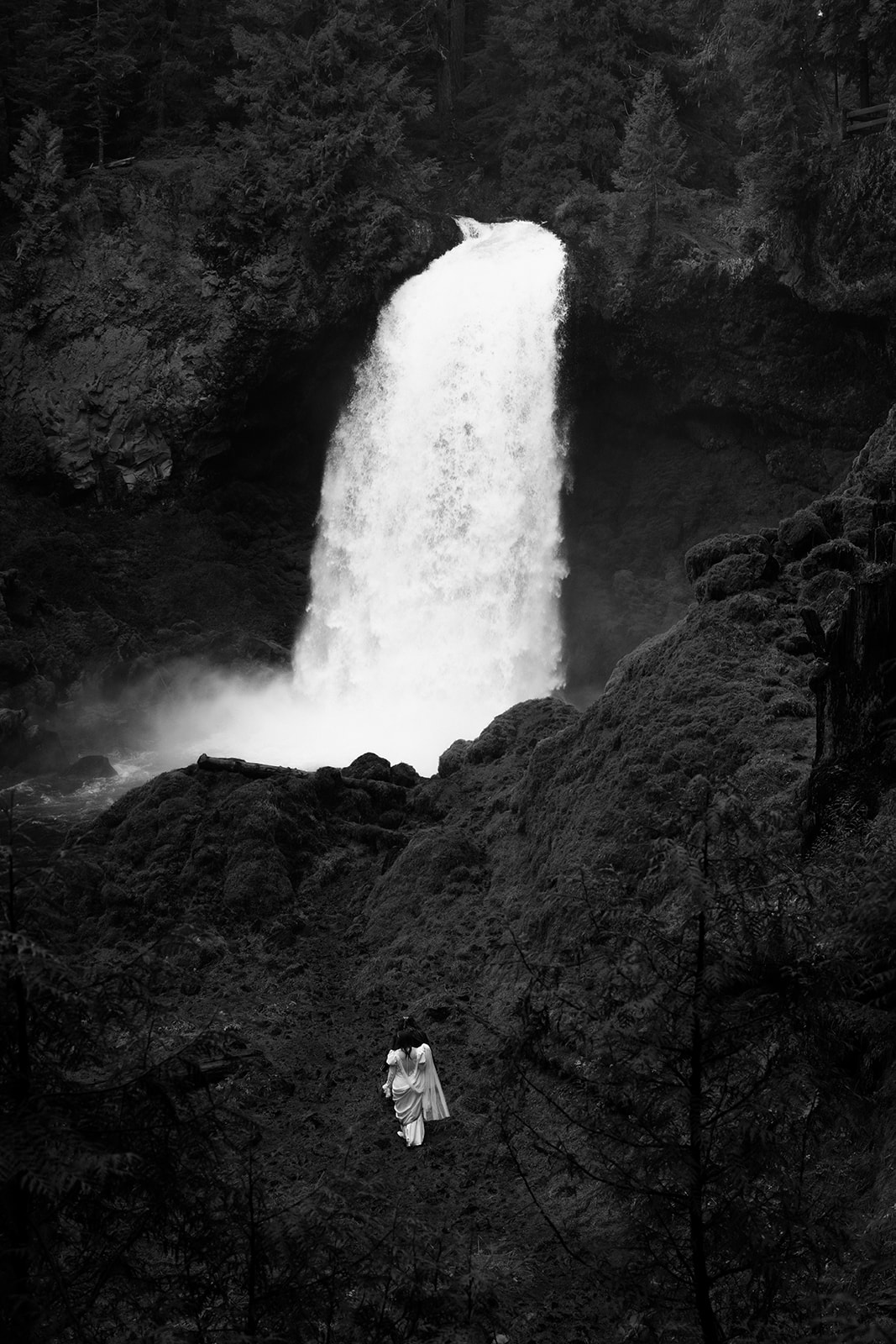 A black and white photo of a giant waterfall with a couple looking small in front of it for a Sahalie Falls elopement. 