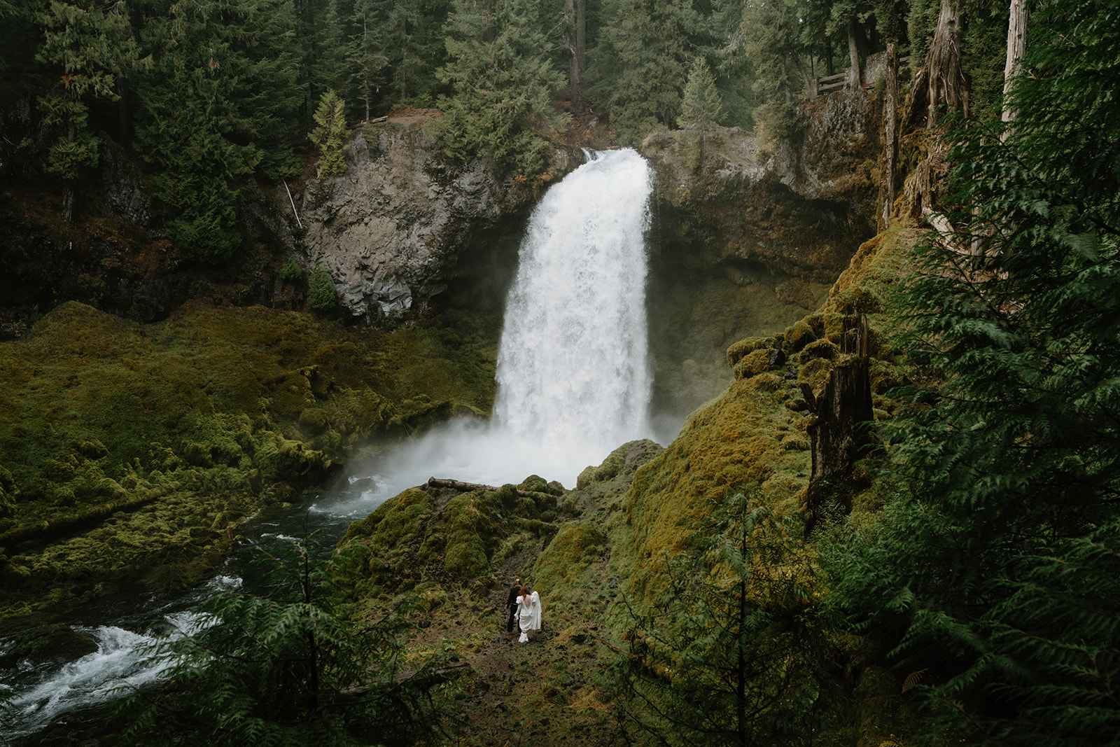 A couple in wedding attire stands small in front of a giant waterfall during their Sahalie Falls elopement. 