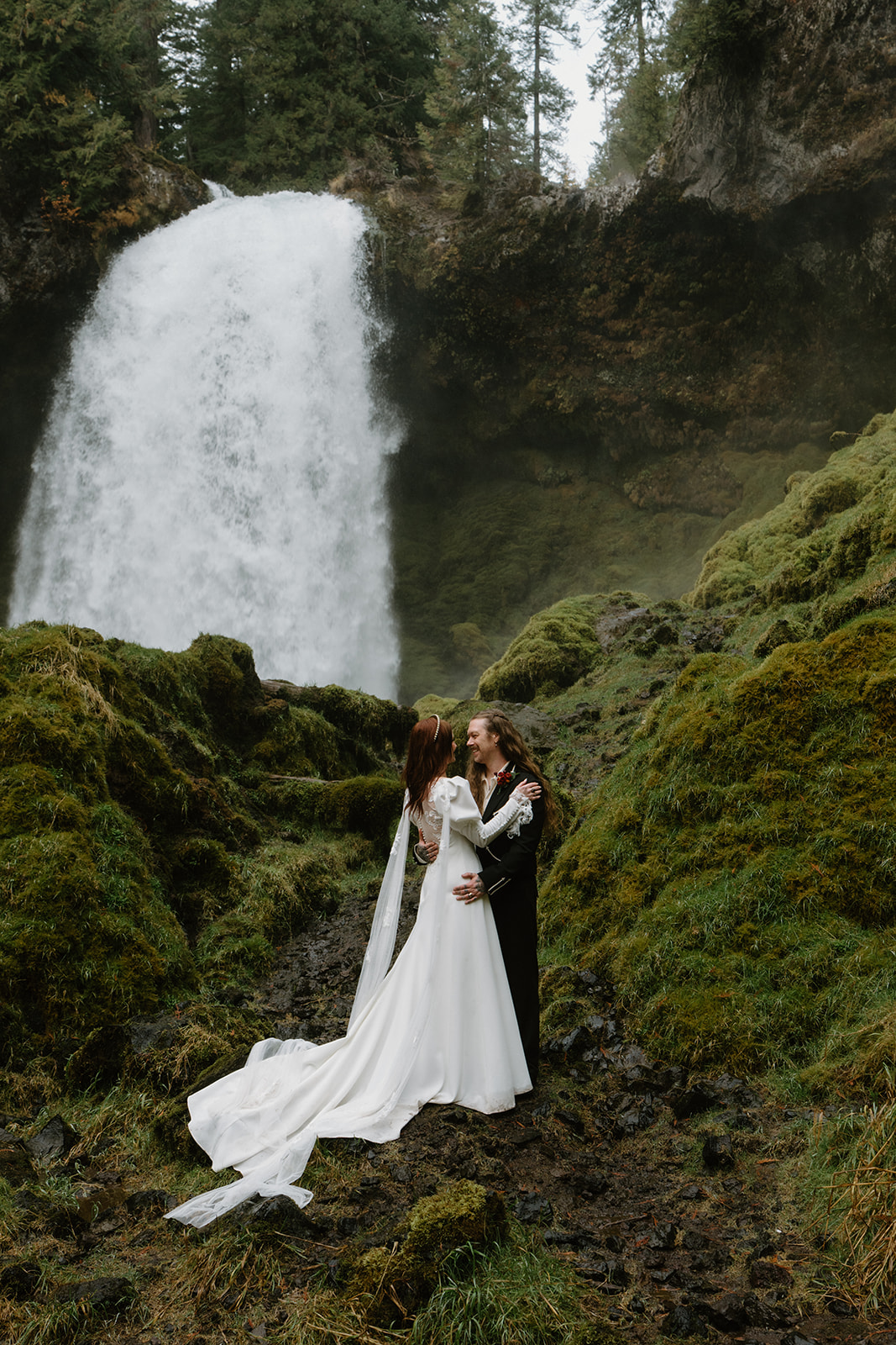 A couple in wedding attire stand in front of a giant waterfall, surrounded by green moss, during their Sahalie Falls elopement. 