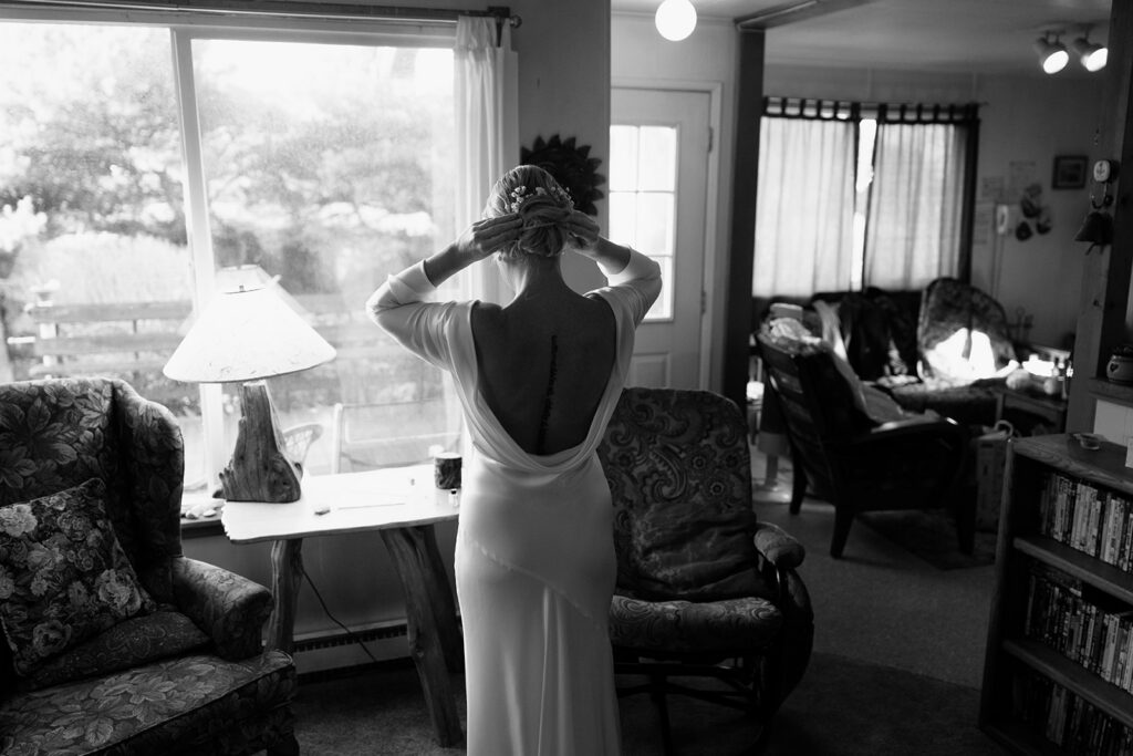 A black and white photo of a bride standing in a living room and putting the final touches on her hair for a Florence, Oregon elopement.