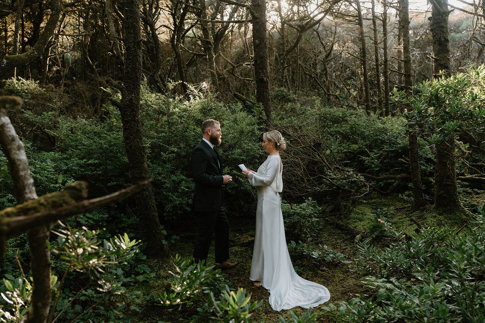A couple in wedding attire stands in the woods. A sunbeam comes through the trees as they exchange vows at their Florence, Oregon elopement.
