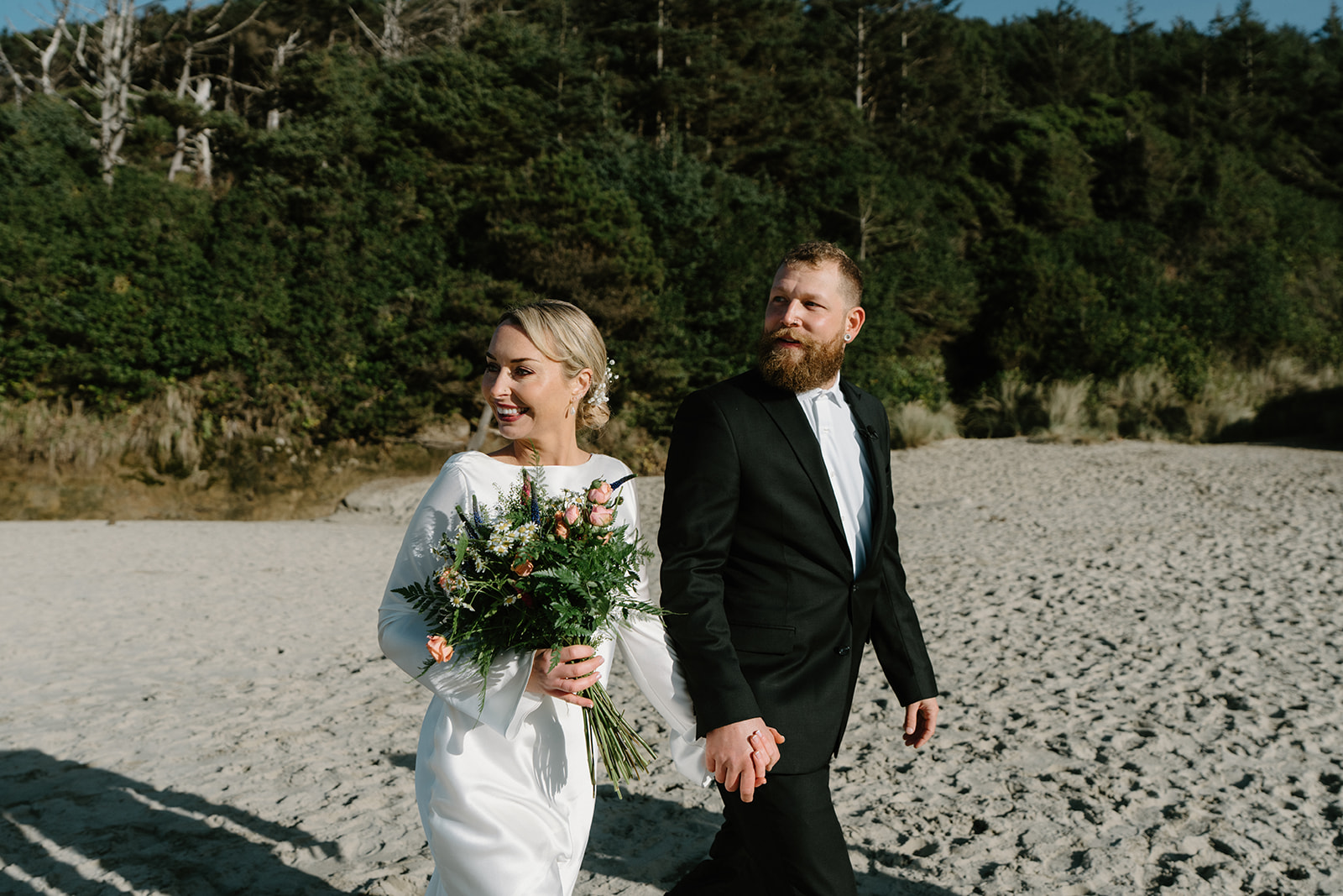 A couple in wedding attire smiles and holds hands and walks on the beach for their Florence, Oregon elopement.