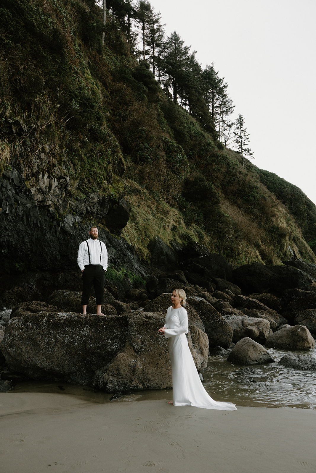 A couple in wedding attire stand near the ocean in front of a rocky black cliff for their Florence, Oregon elopement.