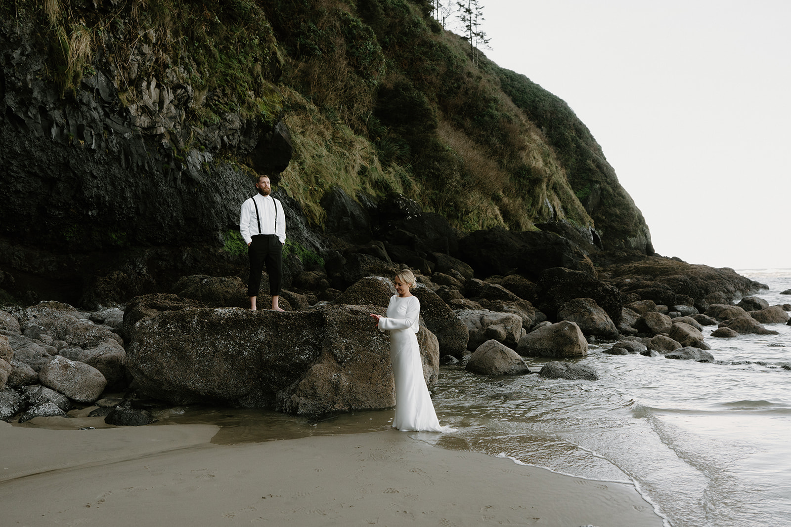 A couple in wedding attire stand near the ocean in front of a rocky black cliff for their Florence, Oregon elopement.